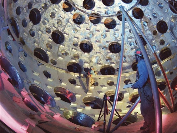 Two people in PPE stand inside of a spherical target chamber where holes provide access for laser beams