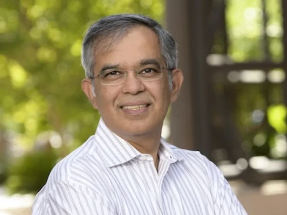 A photograph of Nirav Merchant, standing outside of a sunny day with his arms crossed. He wears glasses and a white shirt.