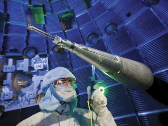 A technician in full cleanroom attire, including a face shield and hood, stands near a long, conical instrument used to position fuel targets inside the National Ignition Facility. The background shows the interior of the target chamber, with glowing blue lighting and metal structures creating a high-tech atmosphere.