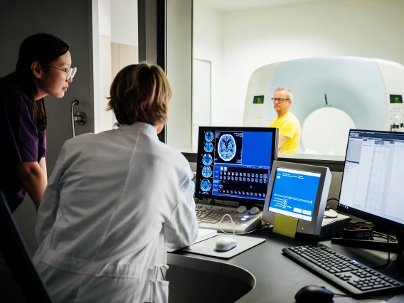A female doctor and a nurse observing medical imaging results on a computer screen in a hospital, while a patient undergoes an imaging procedure. They are located in a control room, with screens displaying medical data and imaging results.