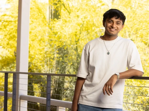 A young man with short dark hair leans casually against a railing, smiling warmly. He wears a white T-shirt, silver wristwatch, and a pendant necklace, with vibrant yellow trees and natural light in the background.