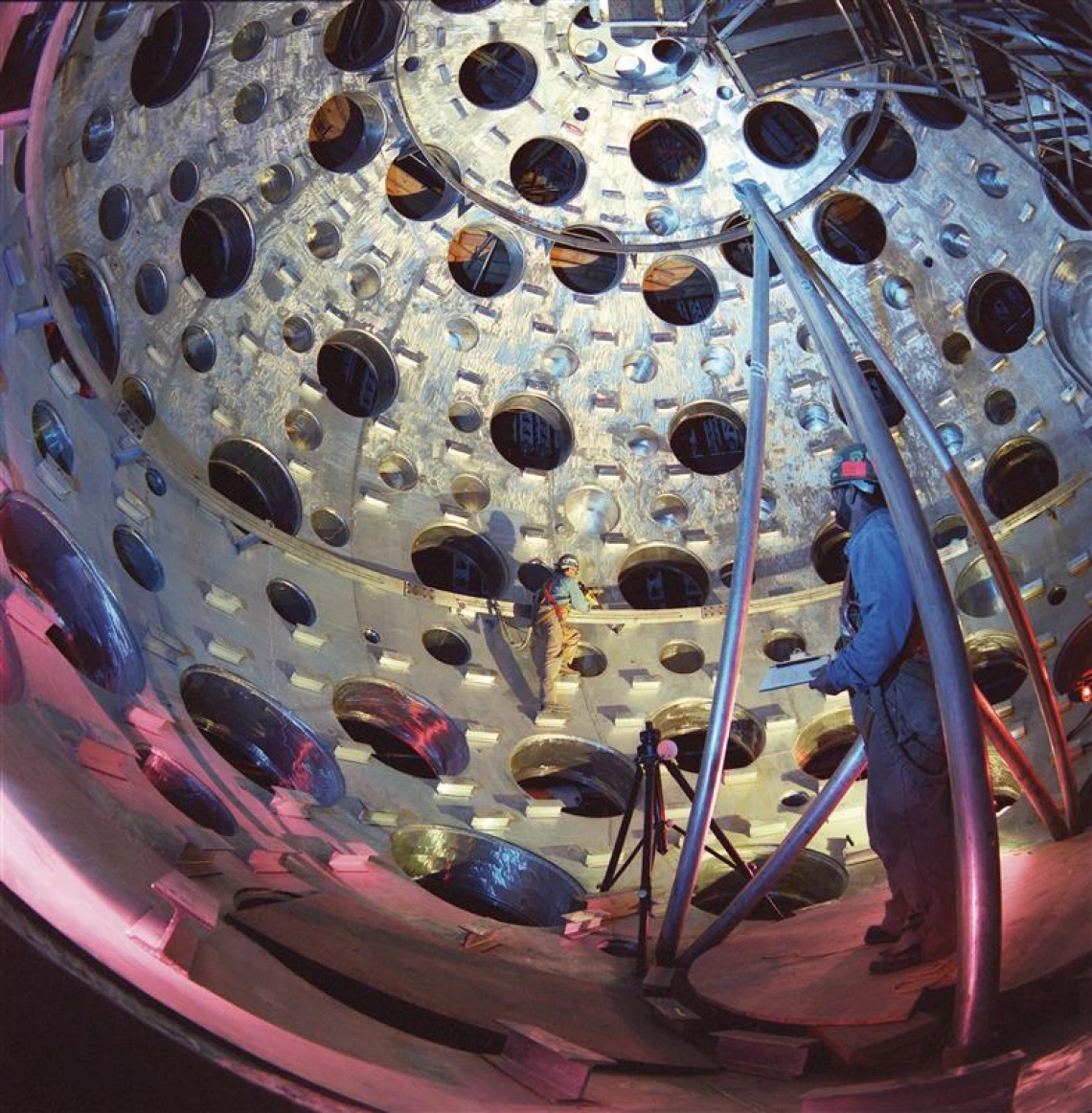 Two people in PPE stand inside of a spherical target chamber where holes provide access for laser beams