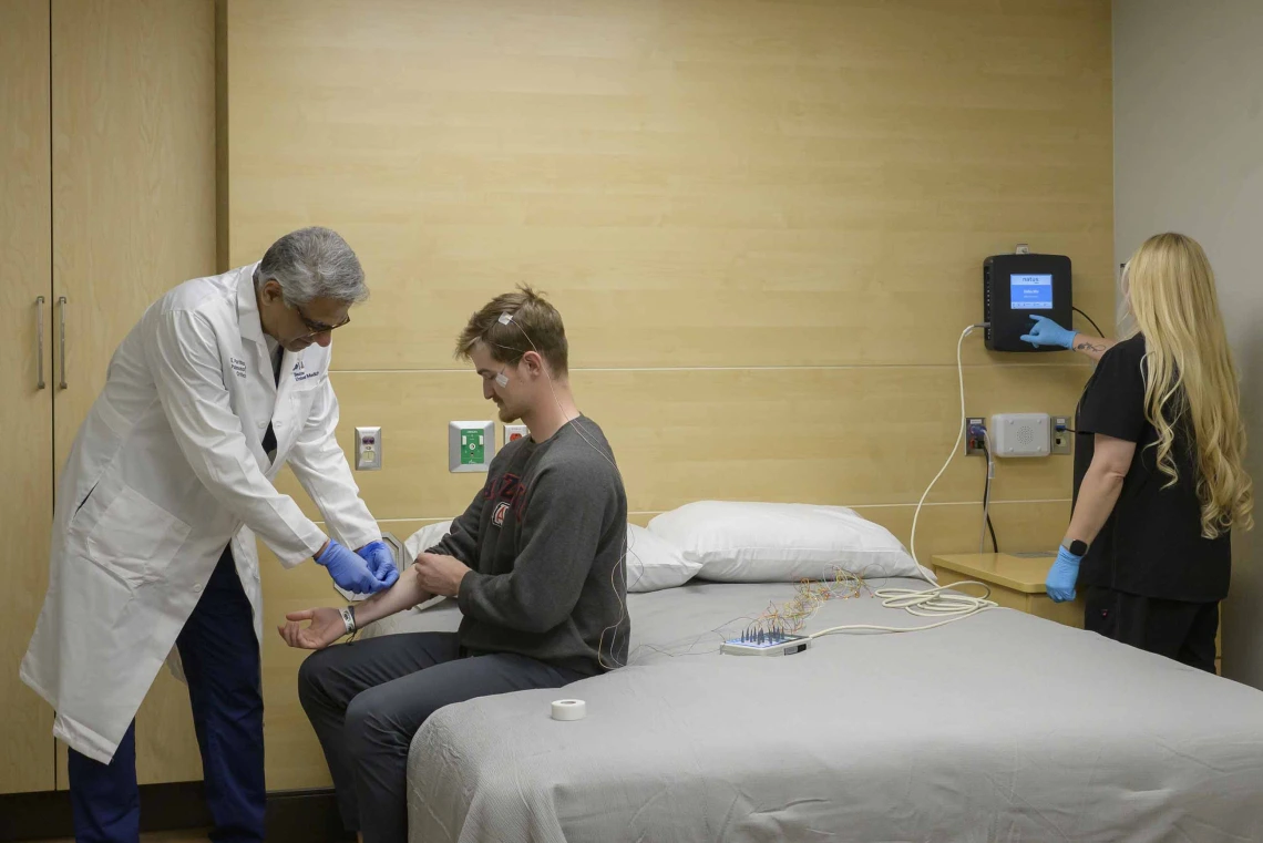 A doctor applies sensors to the arm of a person sitting on a bed in a sleep lab while another person monitors the sensor output on a small digital device on the wall.