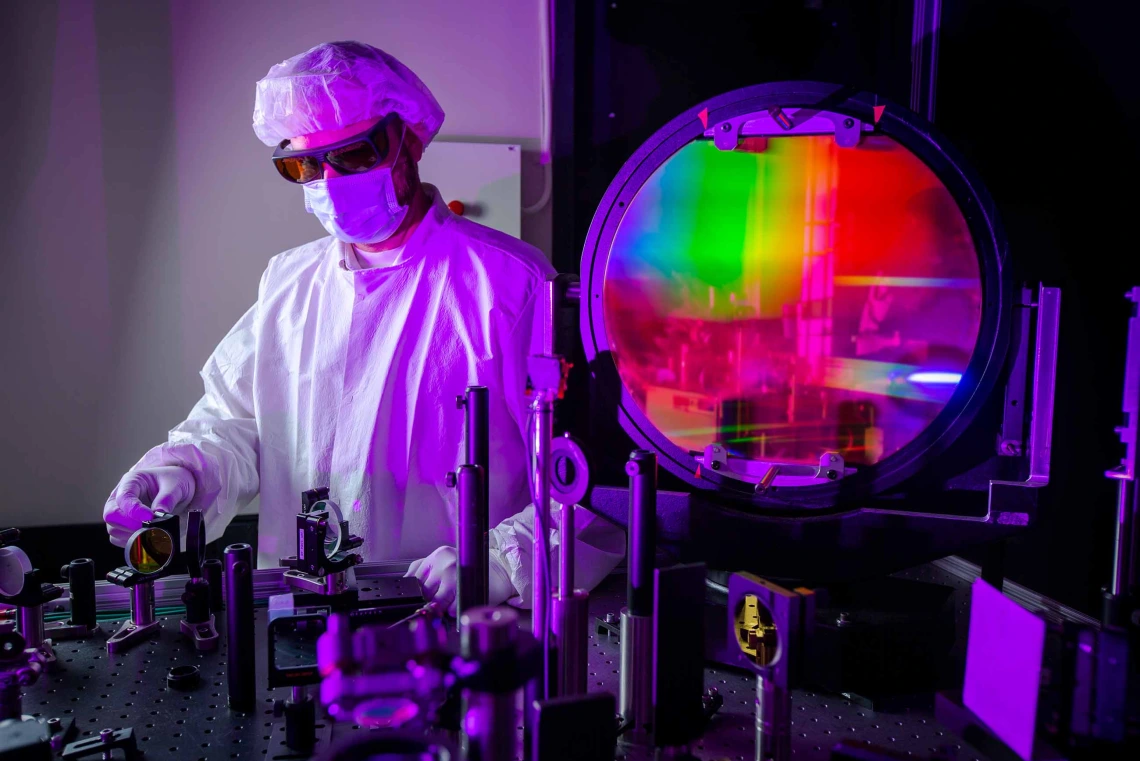 A technician wearing protective clothing and safety glasses adjusts optical components next to a large, colorful laser lens inside a laboratory at Lawrence Livermore National Laboratory. 