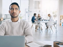 A man sitting at a desk with a laptop and open notebook in front of him looks suspiciously to the side. He is wearing a white knit sweater over a collared shirt. In the background, a group of four people are having a meeting at a table in a brightly lit, modern office space.